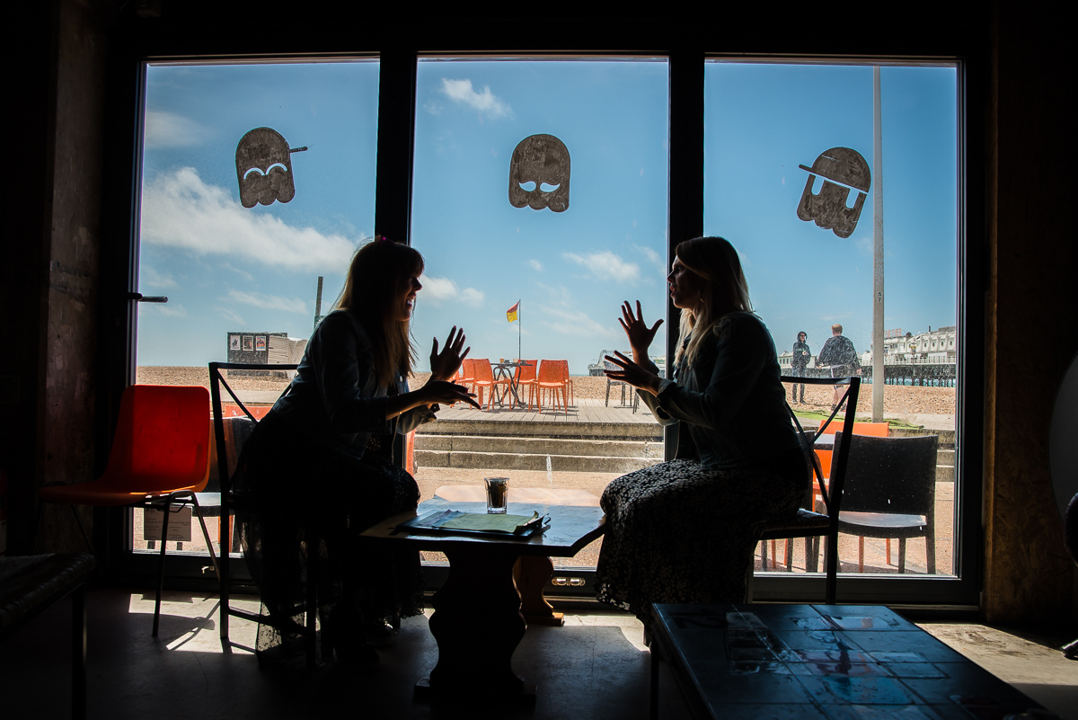 colour image of 2 ladies signing in a cafe against the window with the beach outside