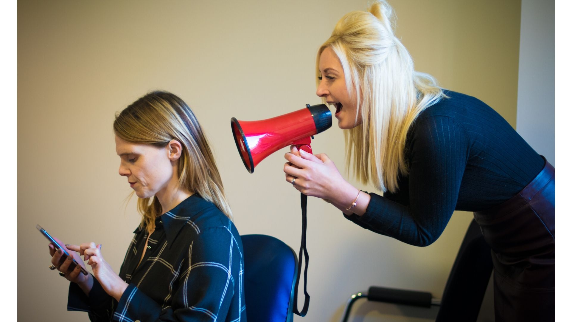 Jennie is sat with her hearing aid visible looking at her phone, a lady is stood behind her shouting into a megaphone.