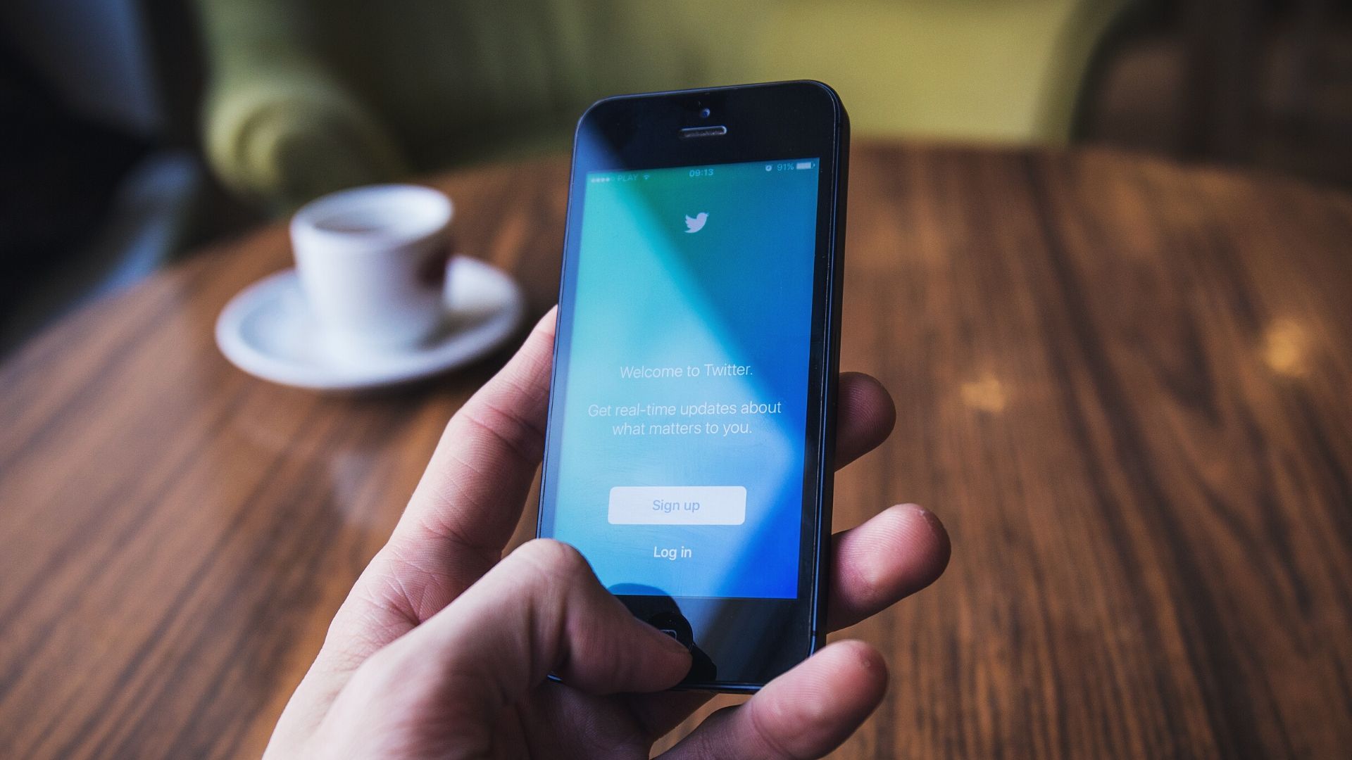 Learn how to make twitter content accessible to visually impaired people.A man's hand holding a mobile phone with Twitter on the screen. A brown table and white coffee cup are in the background.