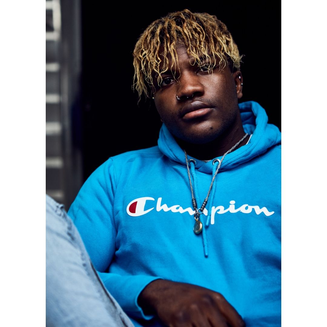Brinston Tchana looks straight at the camera, he is wearing a bright sky blue hoodie and has blonde streaks in his hair.