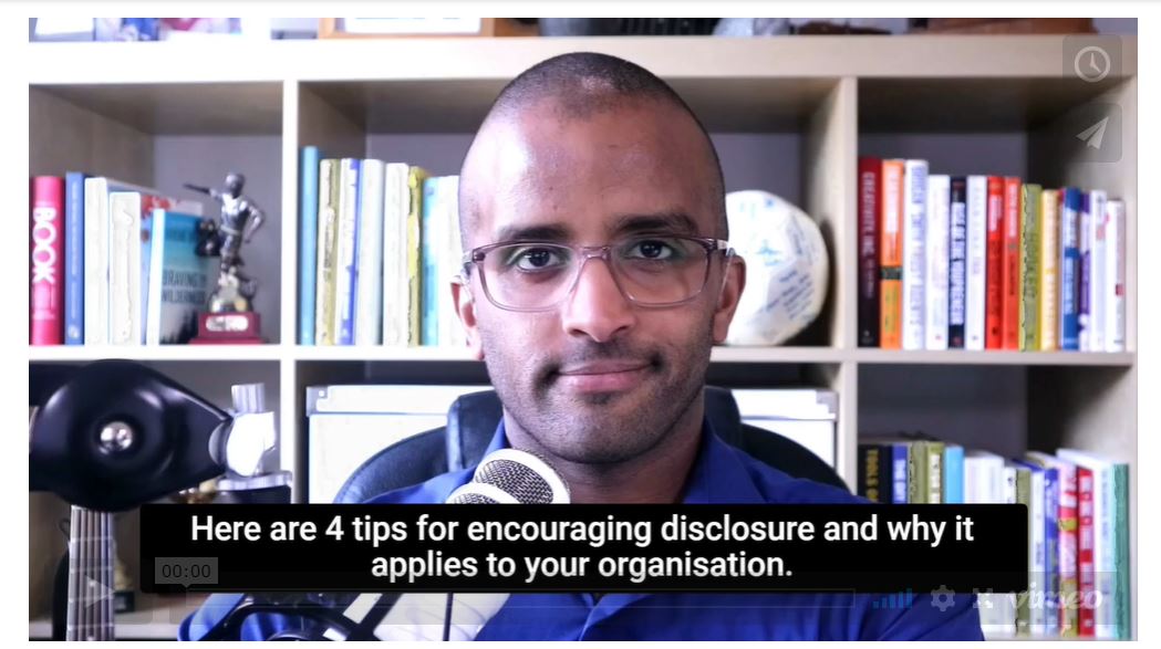 Ahmed is looking at the camera with a microphone in front of him and a bookshelf behind him