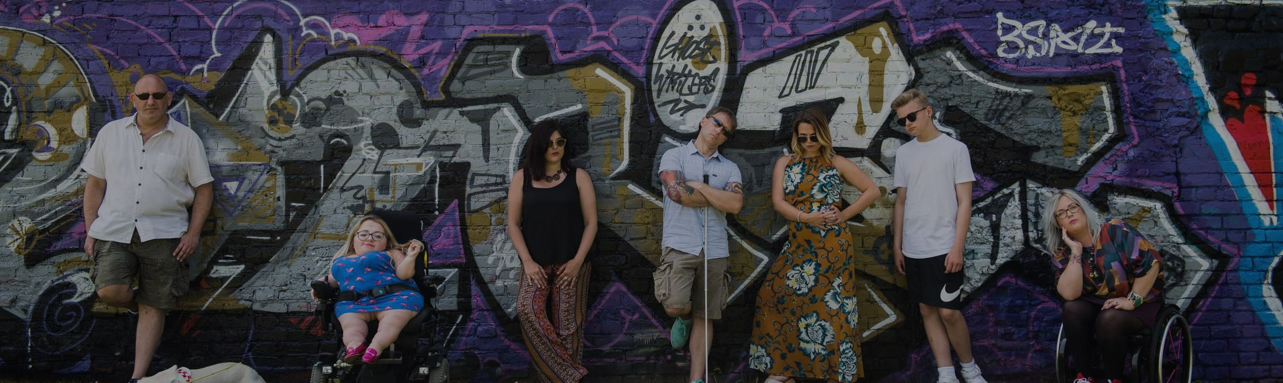 7 members of the Enhance the UK team moodily posing against a graffiti backdrop. One man has a guide dog, another a white cane , one female has restricted growth and is sat in a powerchair and another is in a manual wheelchair with pink spokes.